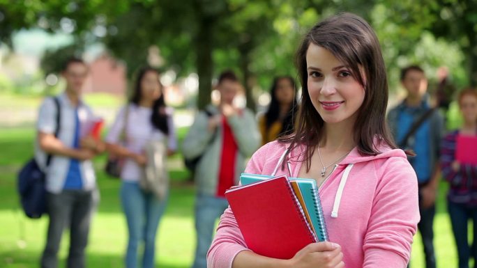 在大学校园的草地上，一个学生对着镜头微笑，她的朋友站在她身后