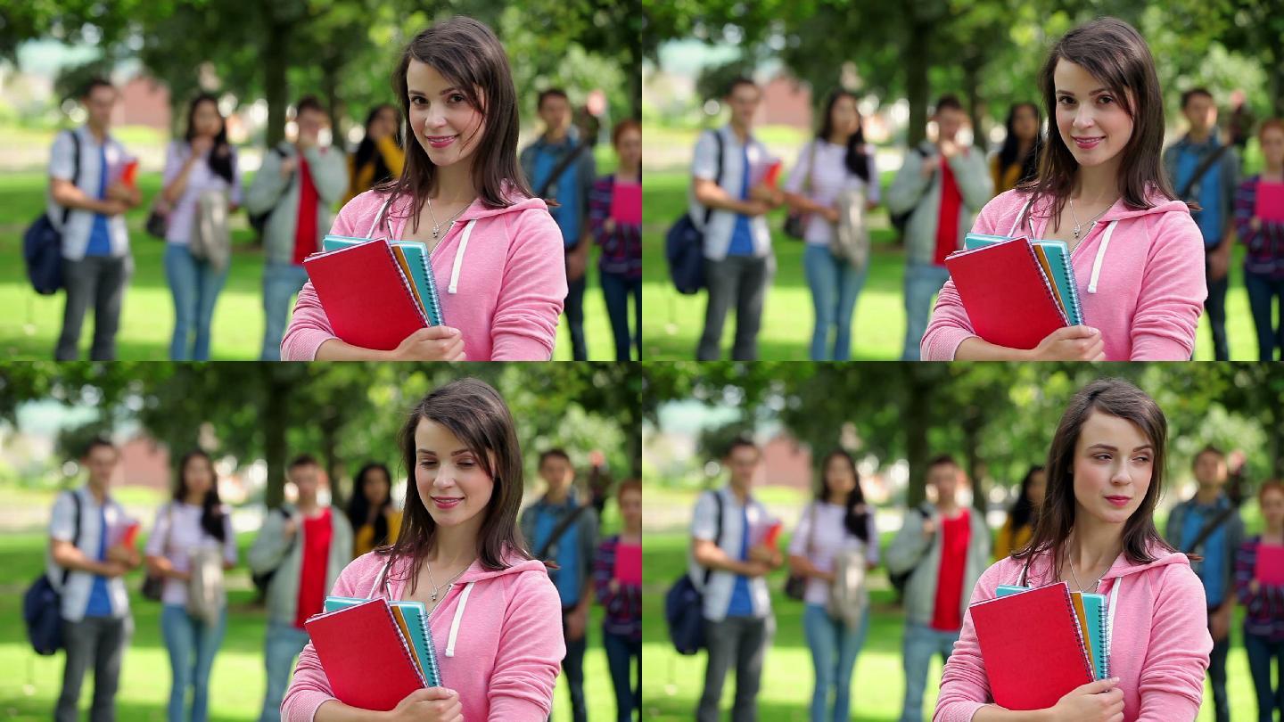 在大学校园的草地上，一个学生对着镜头微笑，她的朋友站在她身后