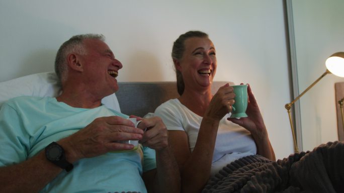 一对白人老夫妇在家里的床上喝着咖啡杯大笑