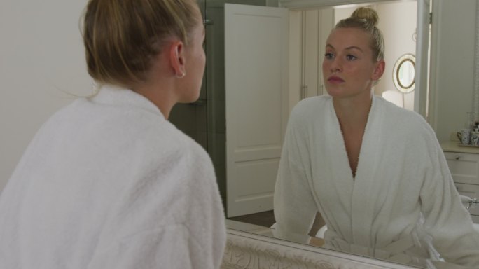 穿着浴衣的白人妇女在浴室里对着镜子摸自己的脸