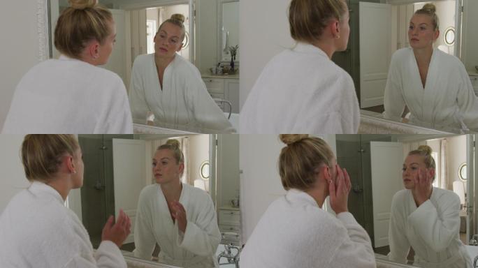 穿着浴衣的白人妇女在浴室里对着镜子摸自己的脸