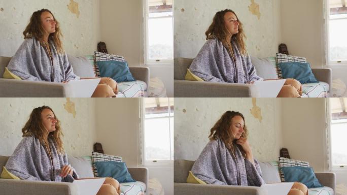 在阳光明媚的小屋客厅里，穿着毯子坐在沙发上使用笔记本电脑的深思熟虑的白人妇女