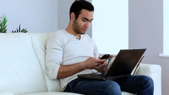 一名男子坐在沙发上用他的智能手机和笔记本电脑