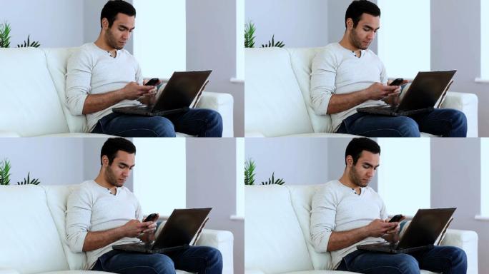 一名男子坐在沙发上用他的智能手机和笔记本电脑