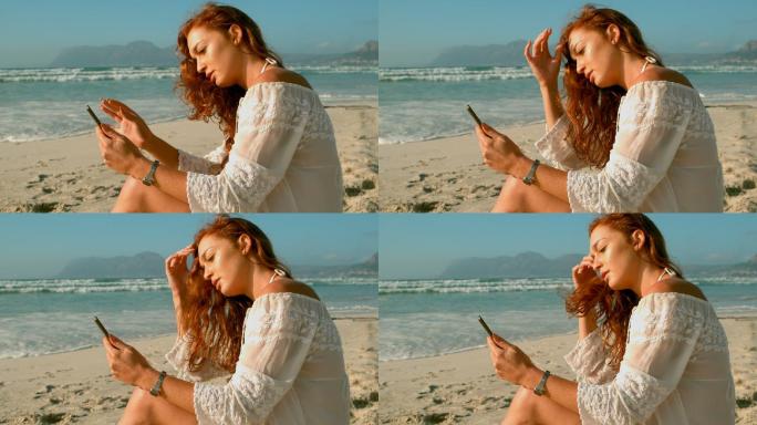 一个白人女人坐在沙滩上玩手机，享受4k的阳光