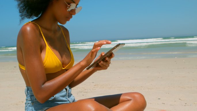 美丽年轻的非洲裔美国女人在沙滩上使用数码平板电脑在4k的阳光下