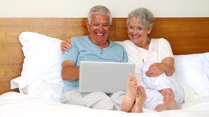 一对老年夫妇坐在床上，在卧室里用笔记本电脑