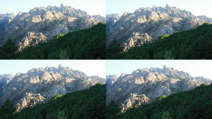 秃顶的石白色滴水石形状的山，后面是茂密的蔓生冷杉和落叶树木的绿色森林。太阳照在山上。