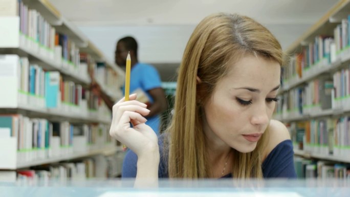 白人女大学生在图书馆看书，背景是一个非裔美国人从书架上拿书。推拉铅球