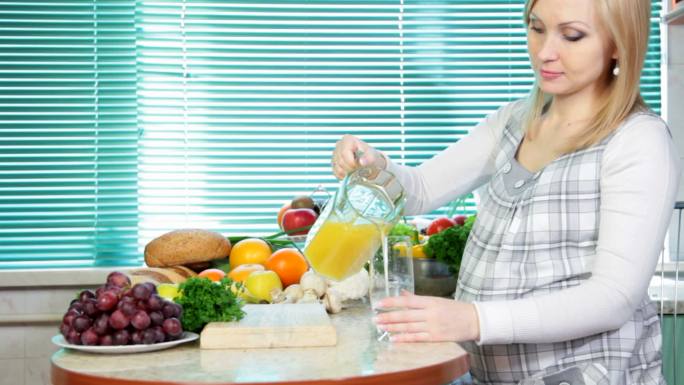 孕妇在厨房里倒橙汁，旁边有很多蔬菜和水果