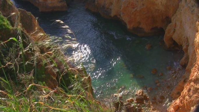 从一块高高陡峭的长满草的岩石到一个被裸露的悬崖环绕的岩石海湾；太阳在浅绿色的水中闪闪发光；葡萄牙阿尔
