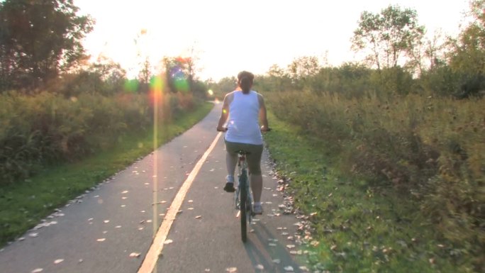 一名年轻女子骑着自行车在一条铺好的草原小道上，穿过森林，来到日落时分。