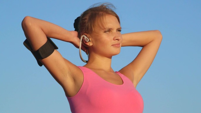 戴着耳机和运动智能手机臂章的健身女性在训练中眺望蓝天