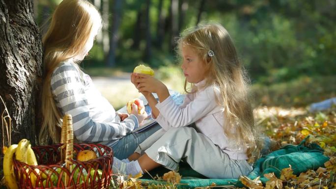 两个金发小女孩在树下享受阳光明媚的秋日，读书和吃苹果