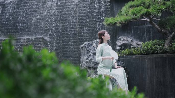 汉服年轻女子摇着扇子坐在园林景观水景墙前