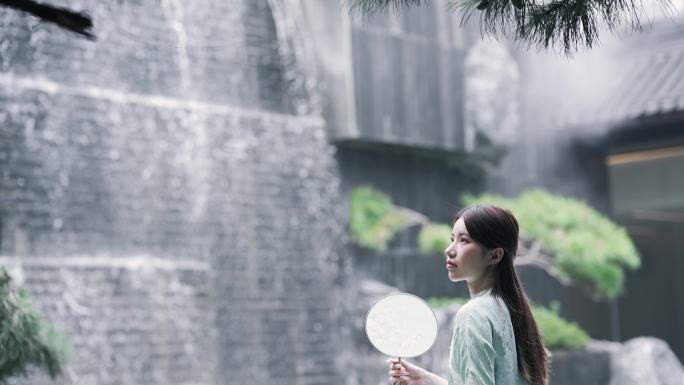 身穿汉服年轻女子摇着团扇坐在水景墙瀑布前