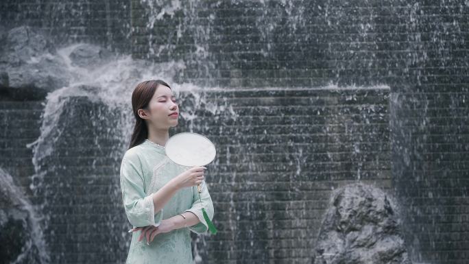 身穿汉服年轻女子在园林景观水景墙前摇扇子