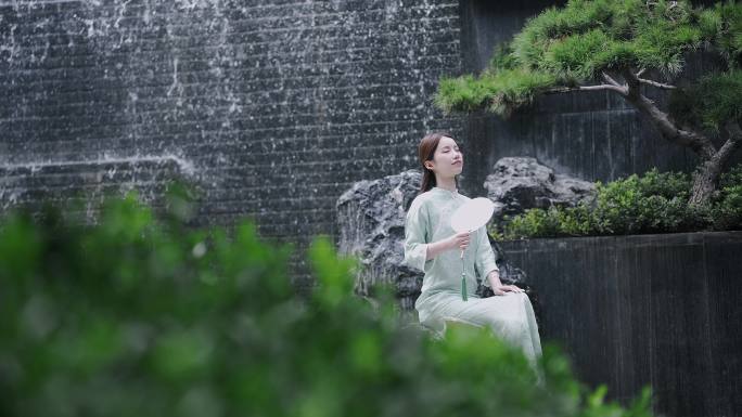 汉服年轻女子摇着扇子坐在园林景观水景墙前