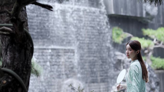 身穿汉服年轻女子摇着团扇坐在水景墙瀑布前