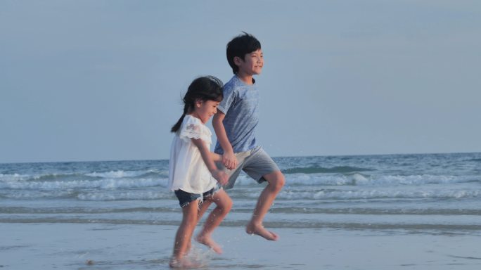 幸福两个4-8岁的亚洲男孩和女孩手牵着手，在沙滩上沿着海边嬉戏。热带夏季假期，活跃的儿童户外活动。帮