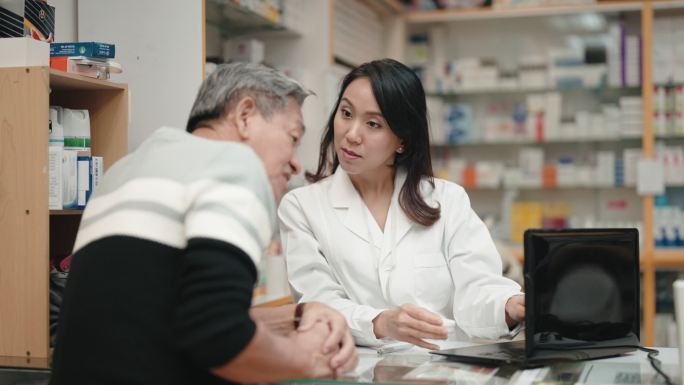 亚洲华人女药剂师在药房柜台向资深男性顾客解释