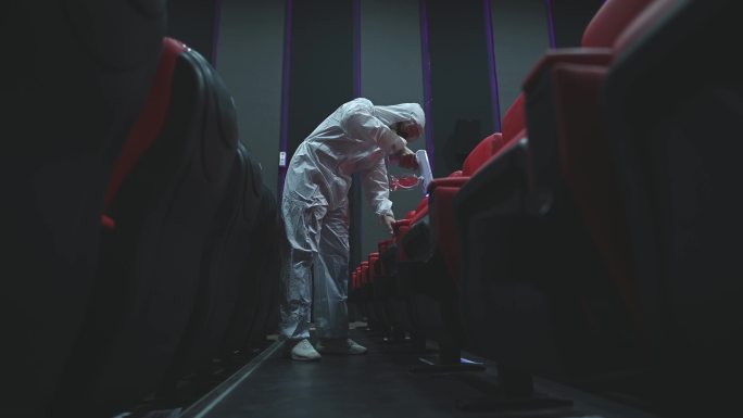 亚裔中国电影清洁工人，戴着口罩、口罩和防护服，在电影放映前在电影院大厅的椅子和地毯上进行消毒