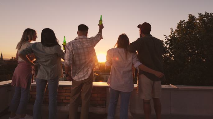夕阳下，朋友们在屋顶上举着啤酒手的背影。