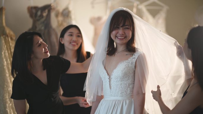 亚洲中国新娘试穿婚纱，伴娘围着她，在婚纱店欣赏婚纱的美丽