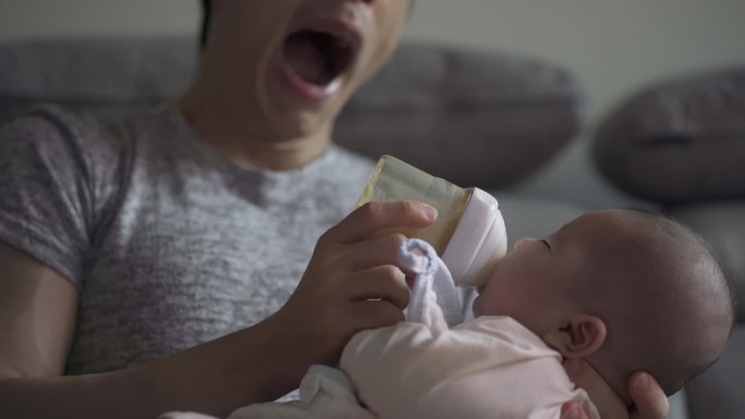 周末，一位疲惫的亚裔中国年轻父亲在客厅打呵欠，用奶瓶喂他的男婴
