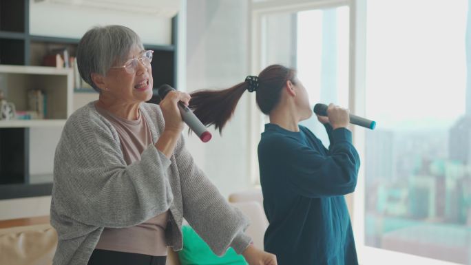 周末休闲活动期间，一名亚裔华裔高级女性在客厅与女儿唱歌、卡拉OK跳舞