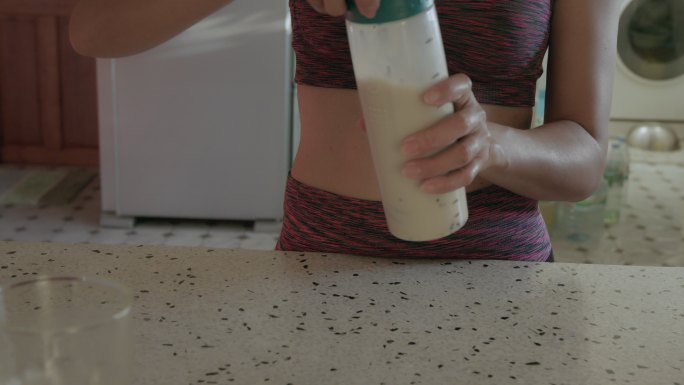 一位穿着运动服的亚洲年轻女子在家里的厨房里用蛋白粉制作奶昔，喝蛋白奶昔。