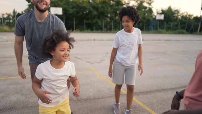一个多种族的继父家庭，一位自豪的父亲，在女儿试图冲入母亲用手制作的想象中的篮球圈时，支持和鼓励女儿