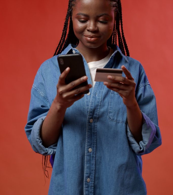 一段垂直视频，一个快乐、有魅力、皮肤黝黑的学生用红色背景的信用卡在智能手机上购买了她非常想要的东西