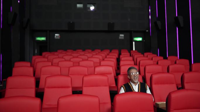 一名留着白胡子的亚裔中国男子坐在空荡荡的电影院里，坐在红色座位上吃东西，欣赏电影。