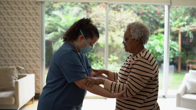 家庭护理人员帮助一名老年妇女站在家中-医生戴着防护面罩