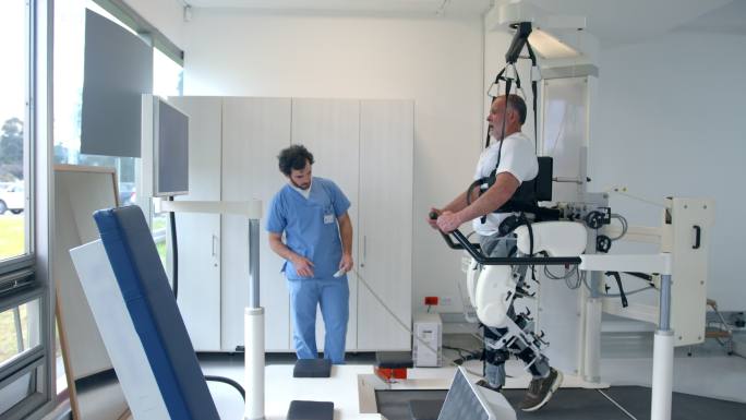 男性治疗师在物理康复治疗期间，在电动外骨骼上向老年患者解释脚的运动