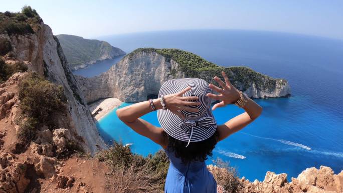4K视频，一名戴着帽子的妇女站在顶部，双臂伸开，看着著名的纳瓦乔沉船海滩，希腊