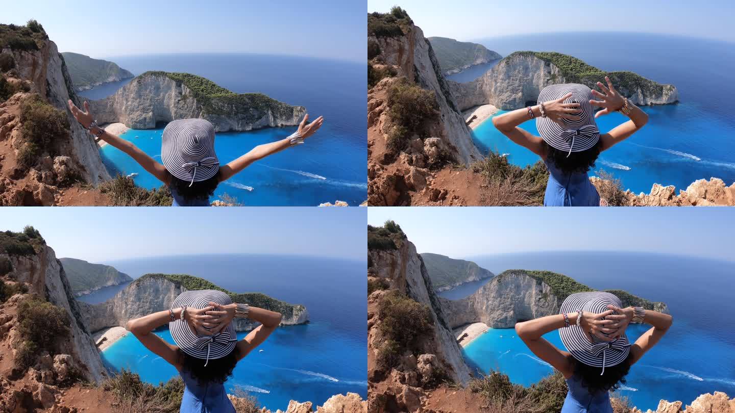 4K视频，一名戴着帽子的妇女站在顶部，双臂伸开，看着著名的纳瓦乔沉船海滩，希腊