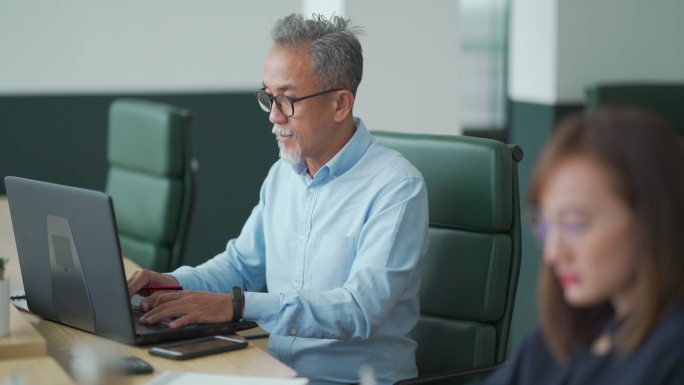 一名亚裔中国工作的高级男子在开放式办公室与同事一起使用笔记本电脑