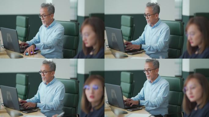 一名亚裔中国工作的高级男子在开放式办公室与同事一起使用笔记本电脑