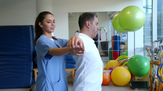 友好的女性治疗师在物理康复治疗期间帮助患者锻炼手臂、肩膀和背部