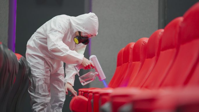 亚裔中国电影清洁工人，戴着口罩、口罩和防护服，在电影放映前在电影院大厅的椅子和地毯上进行消毒