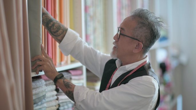 工作室：亚洲中国成熟男装裁缝，脸上留着胡茬，在工作室检查他的织物服装