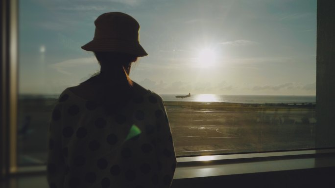 一位年轻的亚洲妇女站在机场的窗口，观察日落时的飞机