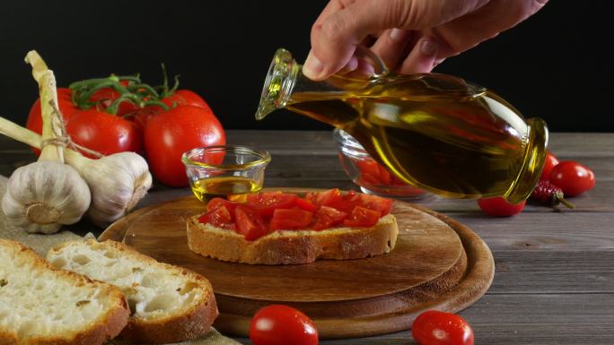 手拿一瓶橄榄油，将特级初榨橄榄油倒入带有西红柿的布氏干酪上，餐桌上摆放着黑色背景的意大利地中海菜肴的