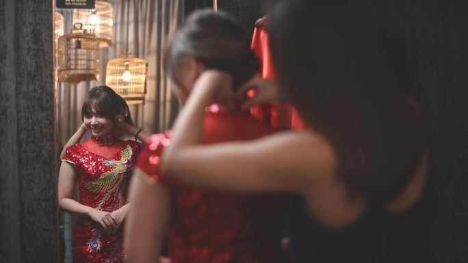 一名亚裔中国妇女在传统精品店试穿中国传统旗袍，在姐姐的帮助下看着镜子