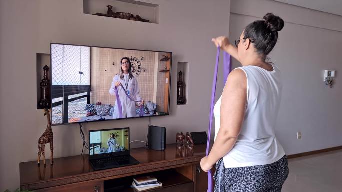 一名成年女性在她的起居室里观看和模仿由物理治疗师在线指导的阻力带练习