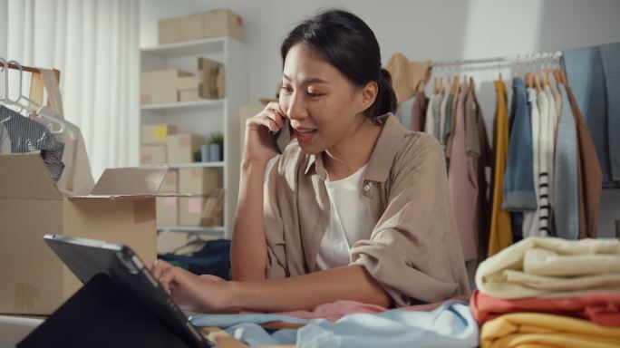 一名年轻的亚洲小企业主妇女正在使用手机接收订单，并在家中的平板电脑上查看库存。
