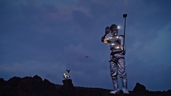 自拍出这个世界。宇航员穿着未来派的照明套装，手持自拍杆。无人驾驶飞机在他们上空飞行