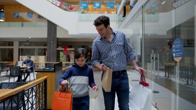 快乐可爱的男孩和他的爸爸在购物中心购物，每个人都提着包，一边愉快地交谈